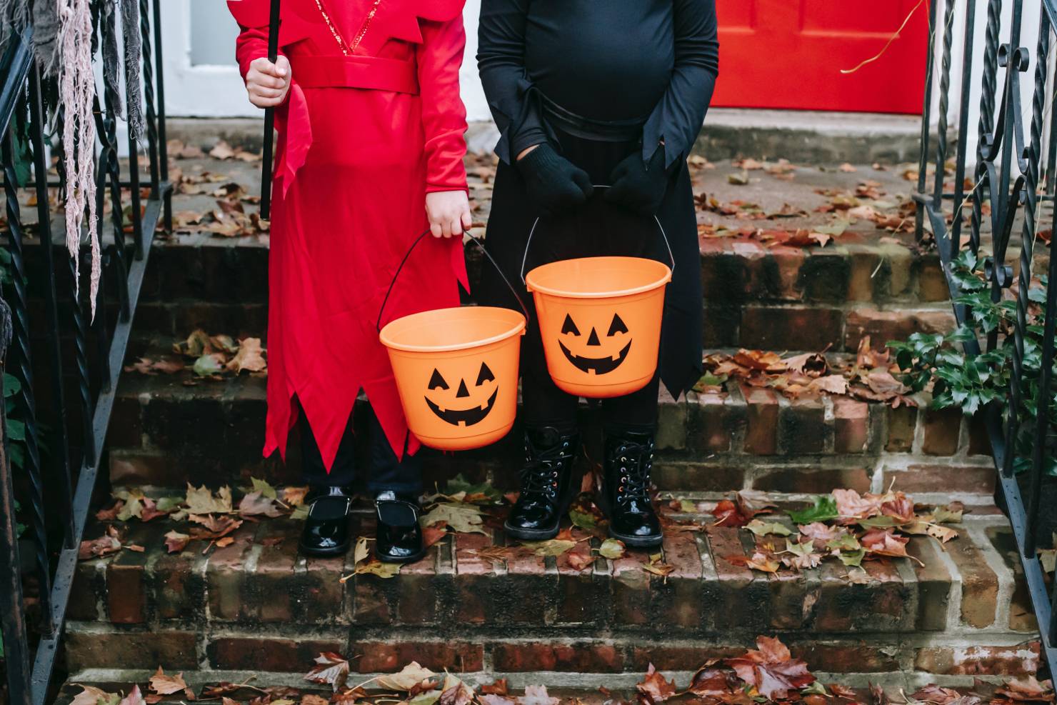 photo of a young girl in a red costume next to a boy wearing a black costume, both holding pumpkin buckets on front door steps, should christians celebrate halloween
