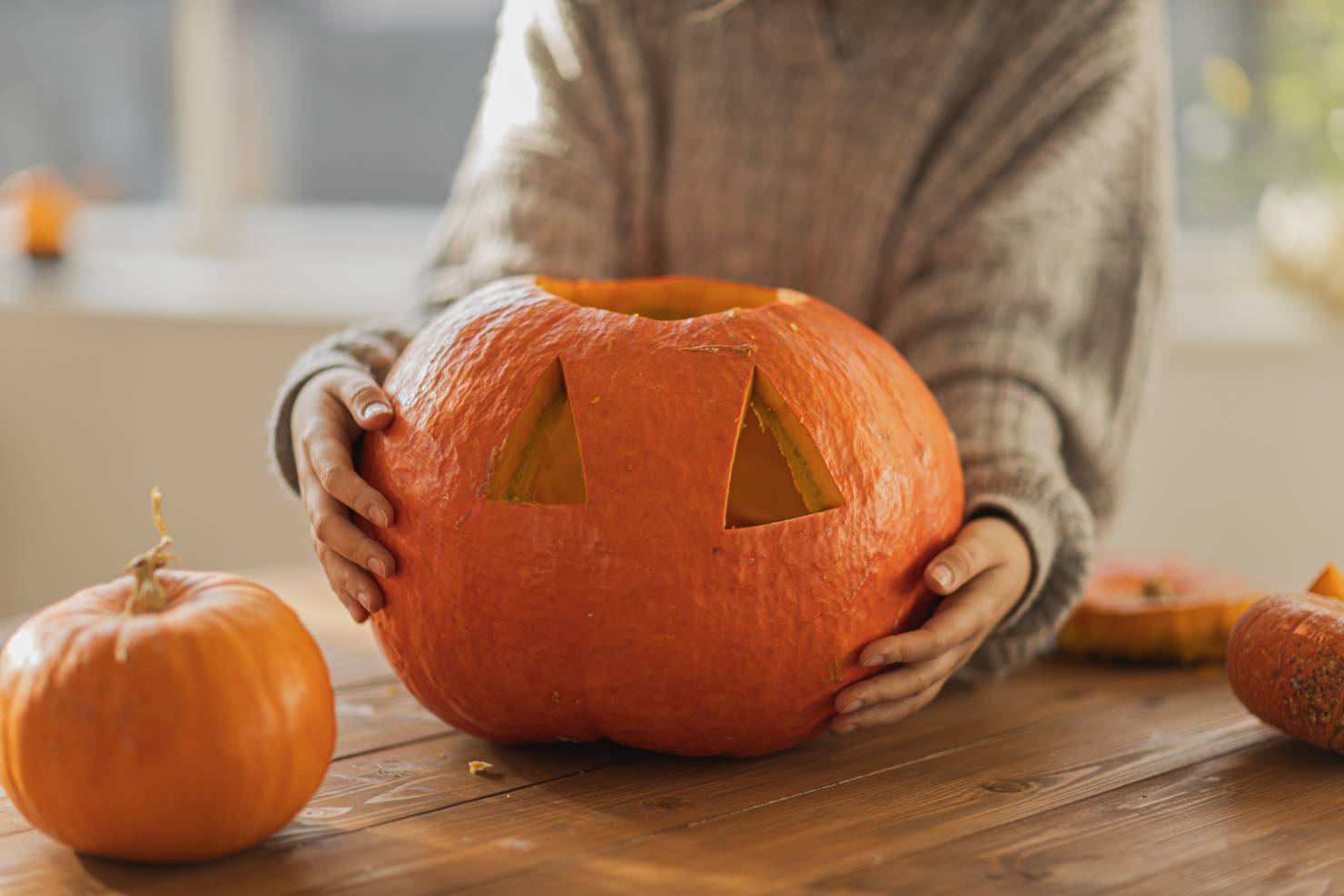 photo of a person wearing a gray sweater, holding a pumpkin with carved out eyes resting on a table