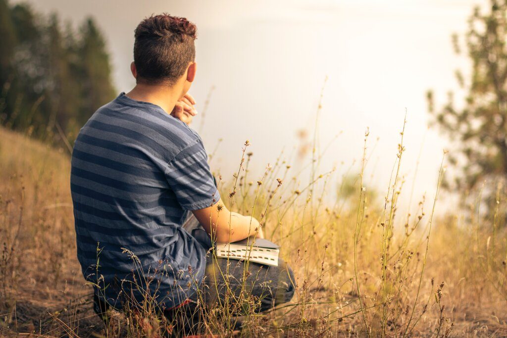 A back view shot of a young men with his bible sitting on a grass field and having quiet time with God 