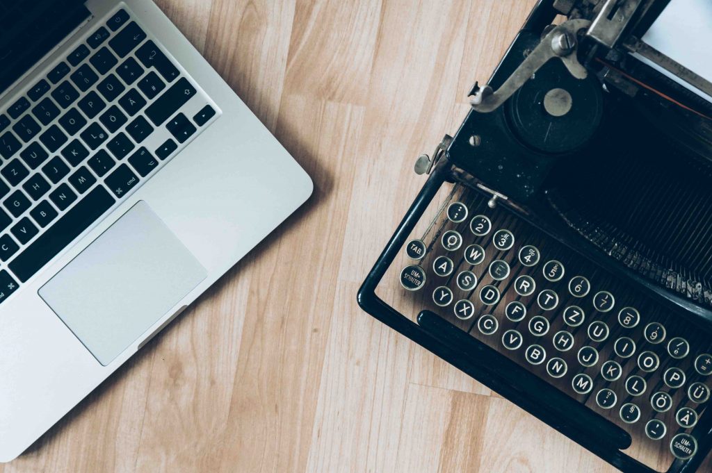 A flat lay image of a macbook and a typewriter on a light wooden table 